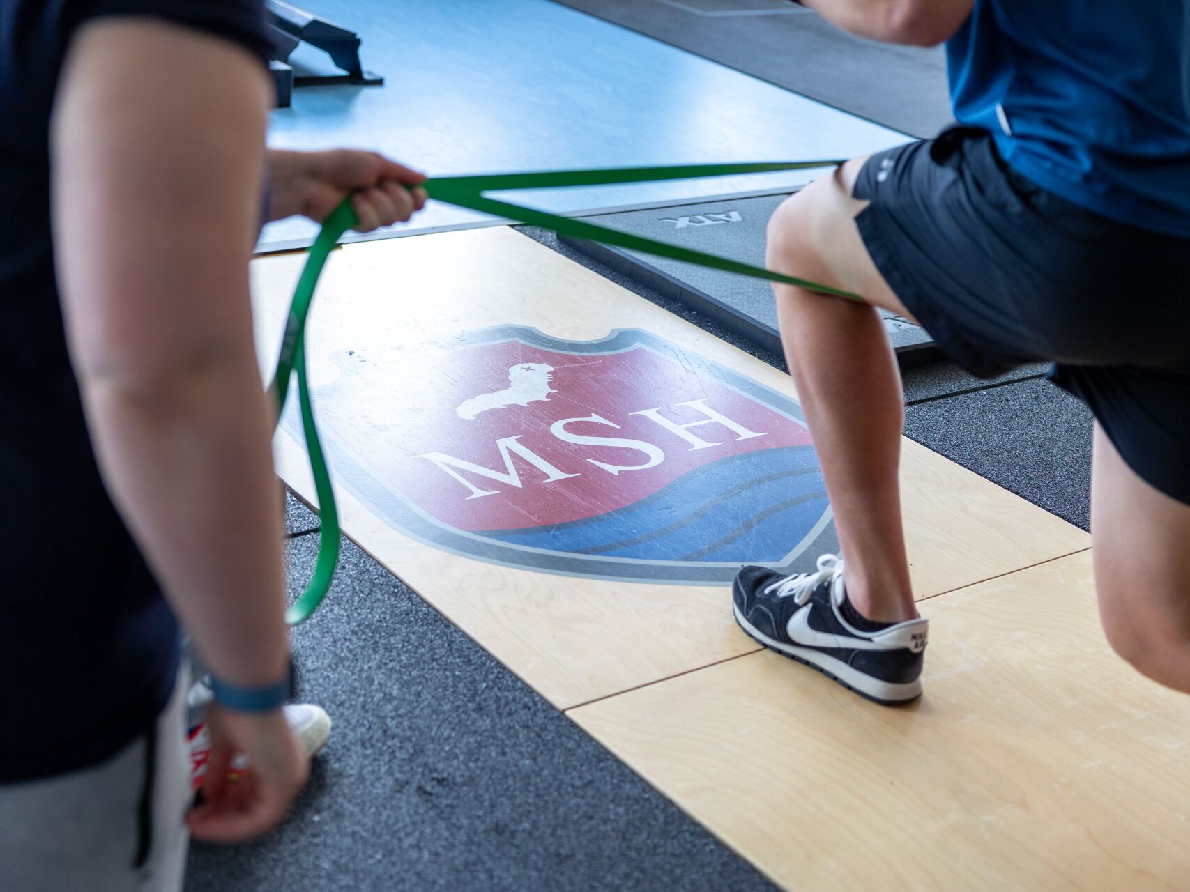 Personen beim Training mit einem Fitnessband in einem Sportraum mit MSH-Logo auf dem Boden.